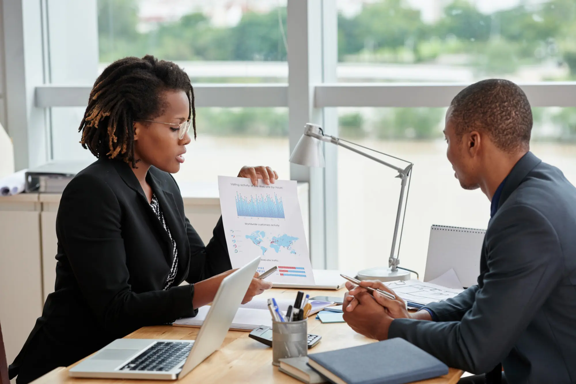 A side view of an African businesswoman presenting a printed visiting-activity chart and worldwide-customer dashboard to her African male coworker across a sunlit desk
